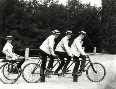 95138 Groepsportret van drie fietsers op een driepersoonsfiets met aanhanger, waarop een vierde persoon, op het ...